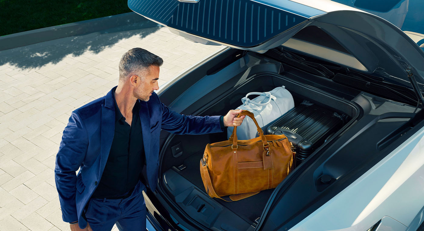 A man in a blue suit places a brown leather bag into the front trunk of a luxury car, conveying a sense of style and travel readiness.